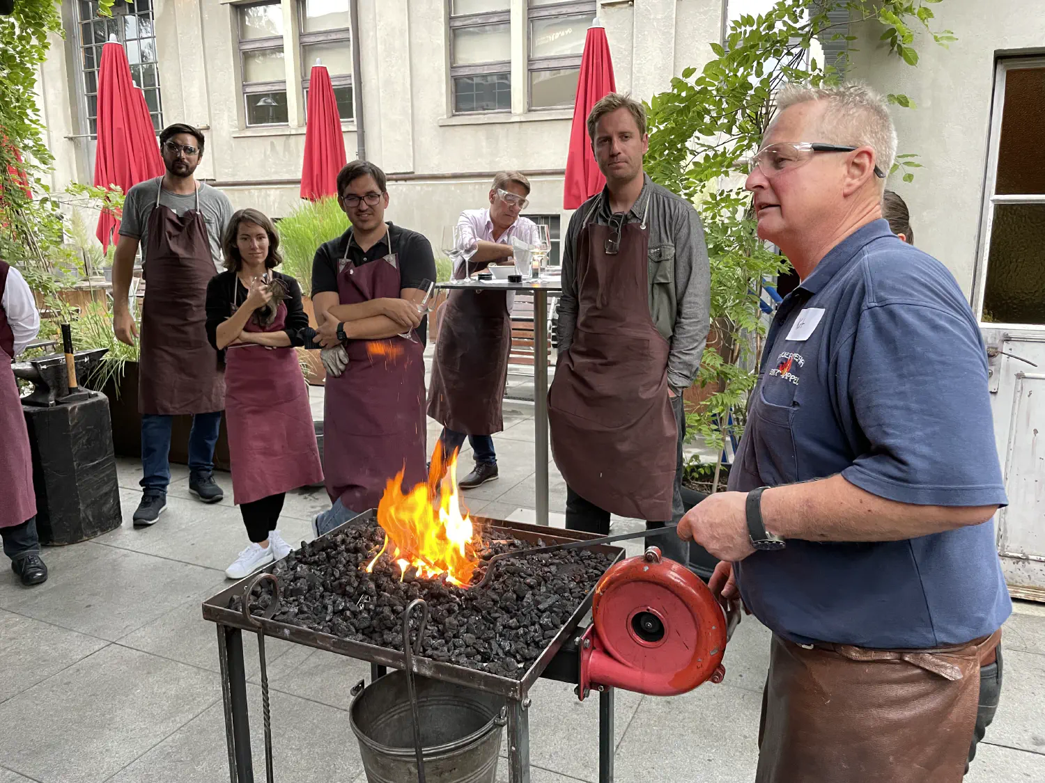 Der Schmied heizt die flammende Kohle mit einem Gebläse an und erklärt wie man das Schmiedegut erhitzt. Die Gäste stehen im Hintergrund und sehen gespannt zu.