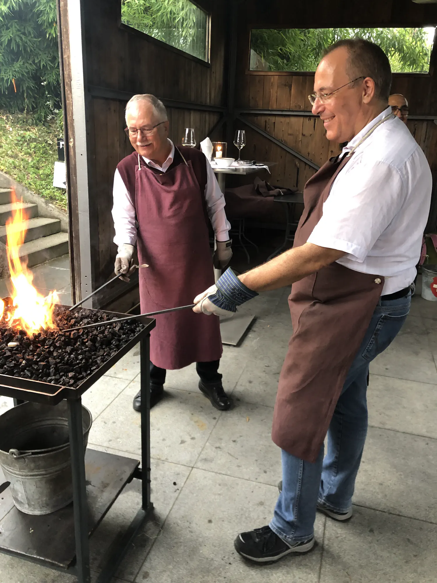 Zwei Personen stehen mit ihren Werkstücken am Kohlefeuer und die Person im Vordergrund lächelt dabei.